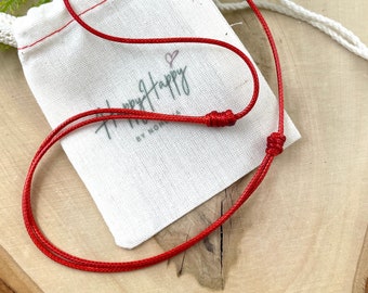 Könnte beinhalten: Eine rote Schnurkette mit einem einfachen Knotenschluss. Die Schnur liegt auf einem weißen Stoffbeutel mit dem Aufdruck "Happy Happy".