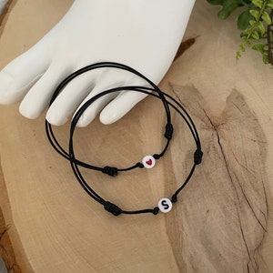 May include: Two black string bracelets with white beads. One bead has a red heart, the other has the letter "S". The bracelets are on a light brown wooden surface.
