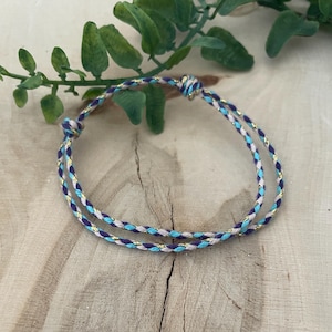 May include: Two braided bracelets with a light blue, purple, and gold color scheme. The bracelets are made of a woven material and are knotted at the ends. The bracelets are displayed on a wooden surface with green foliage in the background.