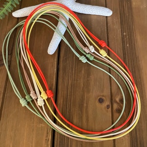Puede incluir: Siete cordones coloridos con extremos anudados. Los cordones son rojo, amarillo, verde claro, beige, verde oscuro y dos tonos de verde claro. Los cordones están dispuestos en círculo sobre una superficie de madera.