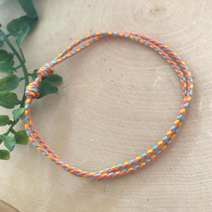 May include: A braided bracelet featuring a vibrant color scheme of orange, yellow, and turquoise. The adjustable bracelet is made of woven cord and is designed to be worn around the wrist. The bracelet is shown on a light brown wooden surface.