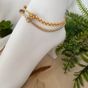May include: Two colorful braided anklets in yellow, pink, blue, and green. The anklets are tied in a knot and are shown on a white mannequin leg.