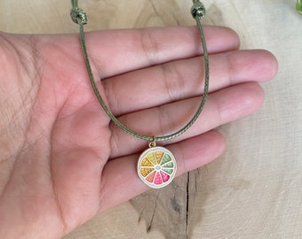 Può includere: Una collana con cordoncino verde con un ciondolo dorato a forma di fetta di agrumi. Il ciondolo è riempito di smalto colorato e ha un contorno bianco.