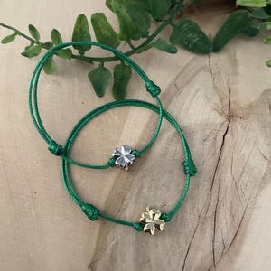 Könnte beinhalten: Zwei grüne Armbänder aus Schnur mit vierblättrigen Kleeblatt-Anhängern. Ein Anhänger ist silberfarben und der andere ist golden.