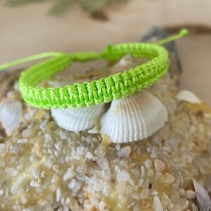 May include: A neon green macrame bracelet. The bracelet is displayed on a bed of sand and seashells. The bracelet is adjustable and appears to be handmade. The bright color of the bracelet contrasts with the neutral tones of the background.