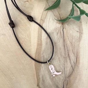 Puede incluir: Un collar de cordón de cuero marrón con un dije de bota rosa y dorada. La bota tiene una estrella dorada en el lateral.