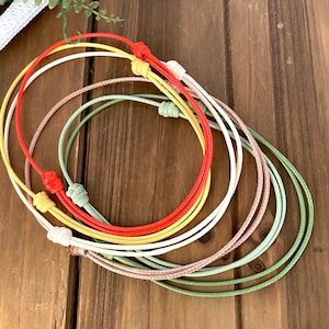 Op de afbeelding: Een set van vijf kleurrijke gevlochten koorden, in tinten groen, roze, wit, geel en rood, zijn aan elkaar geknoopt. De koorden zijn in een cirkel gerangschikt, waardoor een gelaagd effect ontstaat.