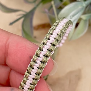 May include: Close-up of a handmade bracelet featuring a woven design. The bracelet combines olive green and light pink cords, creating a textured pattern. The bracelet is held in a hand, showcasing the intricate knotting.