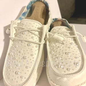 May include: A pair of white canvas slip-on shoes with pearl embellishments. The shoes have a lace-up design and are decorated with small white pearls.