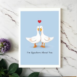 Showcased on a white granite countertop next to vibrant purple flowers, the Ducks in Love Anniversary Card adds a touch of charm and elegance to any special occasion.