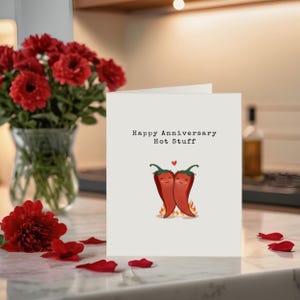 This cute anniversary card, perfect next to flowers, features two adorable hot red peppers hugging with a flame underneath to symbolize fiery love. The message, 'Happy Anniversary Hot Stuff,' adds a romantic touch to your celebration!