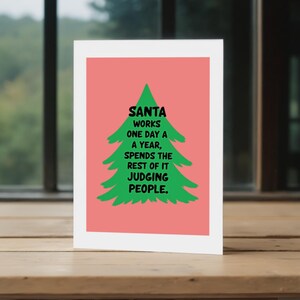 The card has a  background that really pops against the large green Christmas tree silhouette in the center. The tree has a rough, hand-painted appearance with jagged edges, giving it a charming and homemade feel. In bold, black, handwritten-style font, the text "SANTA WORKS ONE DAY A YEAR, SPENDS THE REST OF IT JUDGING PEOPLE". The card is displayed on top of a wooden table.