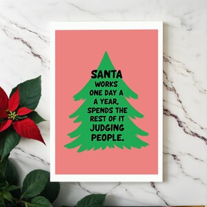The card has a salmon-colored background that really pops against the large green Christmas tree silhouette in the center. The tree has a rough, hand-painted appearance with jagged edges, giving it a charming and homemade feel. In bold, black, handwritten-style font, the text "SANTA WORKS ONE DAY A YEAR, SPENDS THE REST OF IT JUDGING PEOPLE" . This card is displayed on a white granite table.