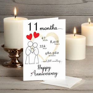 Celebrate your 11 month anniversary with an enchanting card that features a gold heart and intertwined heart balloons held by stick figures. It's presented alongside white candles adding a touch of romance and charm.
