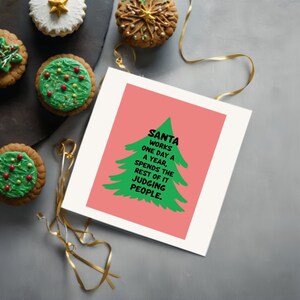 Brighten someone’s holiday with a cheerful Christmas card featuring a playful take on the holiday season! The card has a salmon-colored background that really pops against the large green Christmas tree silhouette in the center. The tree has a rough, hand-painted appearance with jagged edges, giving it a charming and homemade feel. In bold, black, handwritten-style font, the text "SANTA WORKS ONE DAY A YEAR, SPENDS THE REST OF IT JUDGING PEOPLE" . This card is displayed next to festive cupcakes.
