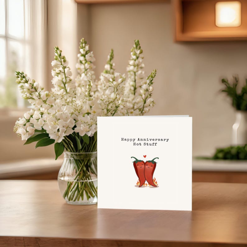 This cute anniversary card, perfect next to white flowers, features 2 adorable hot red peppers hugging with a flame underneath to symbolize fiery love. The message, 'Happy Anniversary Hot Stuff,' adds a romantic touch to your anniversary!