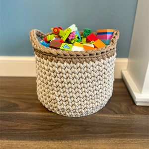 Może przedstawiać: Beżowy koszyk do przechowywania wykonany na szydełku z brązowym górnym brzegiem wypełniony kolorowymi plastikowymi klockami.