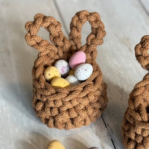 May include: A brown crocheted bunny basket filled with colorful chocolate Easter eggs.  More chocolate eggs are scattered on a white wooden surface.