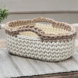 Peut inclure: Un panier crocheté à la main en crème et beige. Le panier a une forme ovale arrondie avec une poignée et un bord tressés de couleur beige contrastante. Le panier est posé sur une surface texturée gris clair.