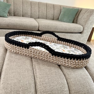 May include: An oval-shaped basket with a thick, beige crocheted base and a black crocheted rim. The interior features a white fabric liner decorated with a colorful pattern of various jungle animals, including giraffes, lions, and tropical foliage.