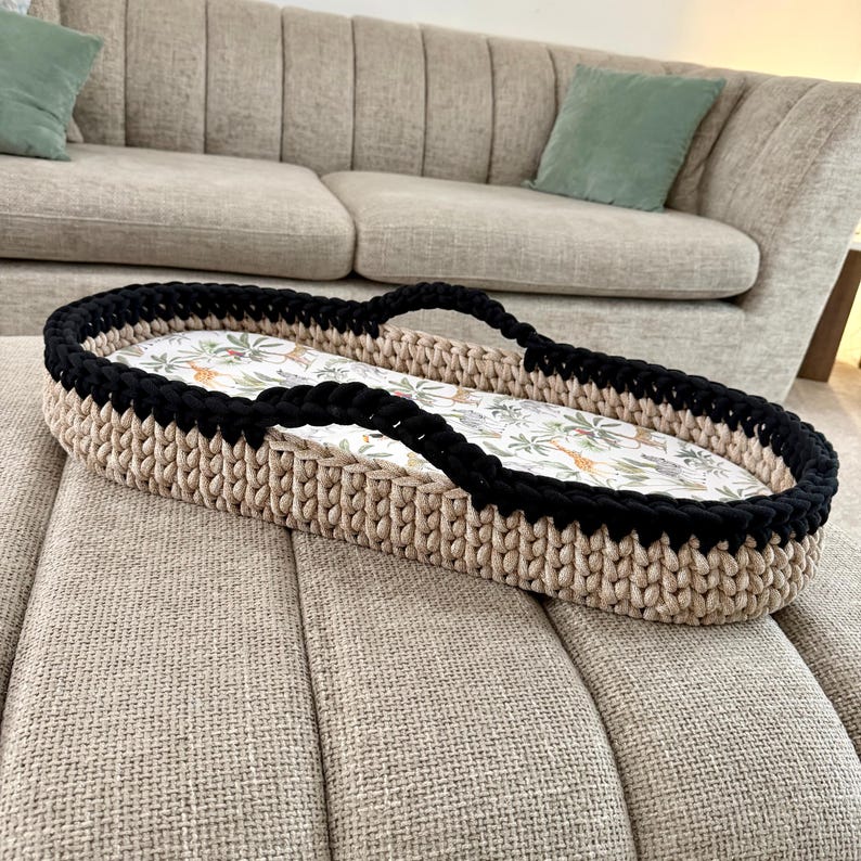 May include: An oval-shaped basket with a thick, beige crocheted base and a black crocheted rim. The interior features a white fabric liner decorated with a colorful pattern of various jungle animals, including giraffes, lions, and tropical foliage.