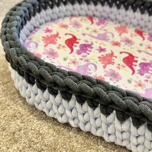May include: An oval-shaped basket featuring a thick, dark gray crocheted rim and a light gray crocheted base. The interior floor of the basket is lined with a fabric pattern displaying red and purple dinosaurs interspersed with small pink flowers.