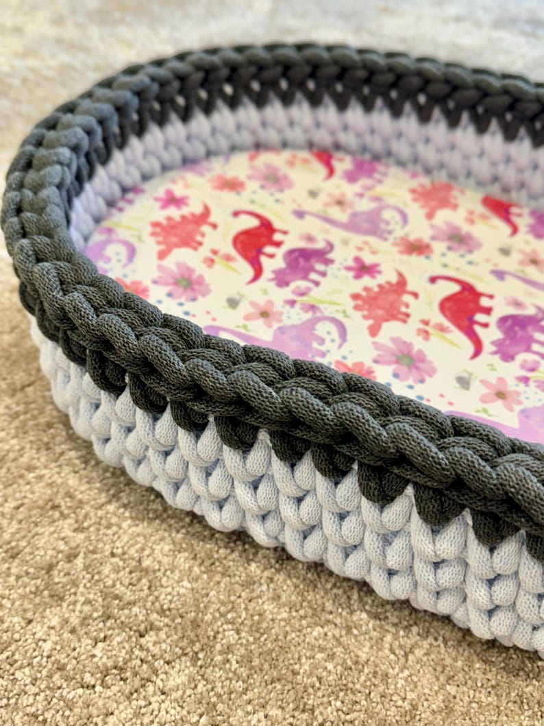 May include: An oval-shaped basket featuring a thick, dark gray crocheted rim and a light gray crocheted base. The interior floor of the basket is lined with a fabric pattern displaying red and purple dinosaurs interspersed with small pink flowers.