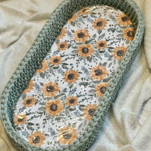 May include: An oval-shaped basket with thick, sage green crocheted walls and a flat base featuring a white fabric liner printed with a repeating pattern of yellow sunflowers, brown centers, and green foliage, covered by a clear plastic film.