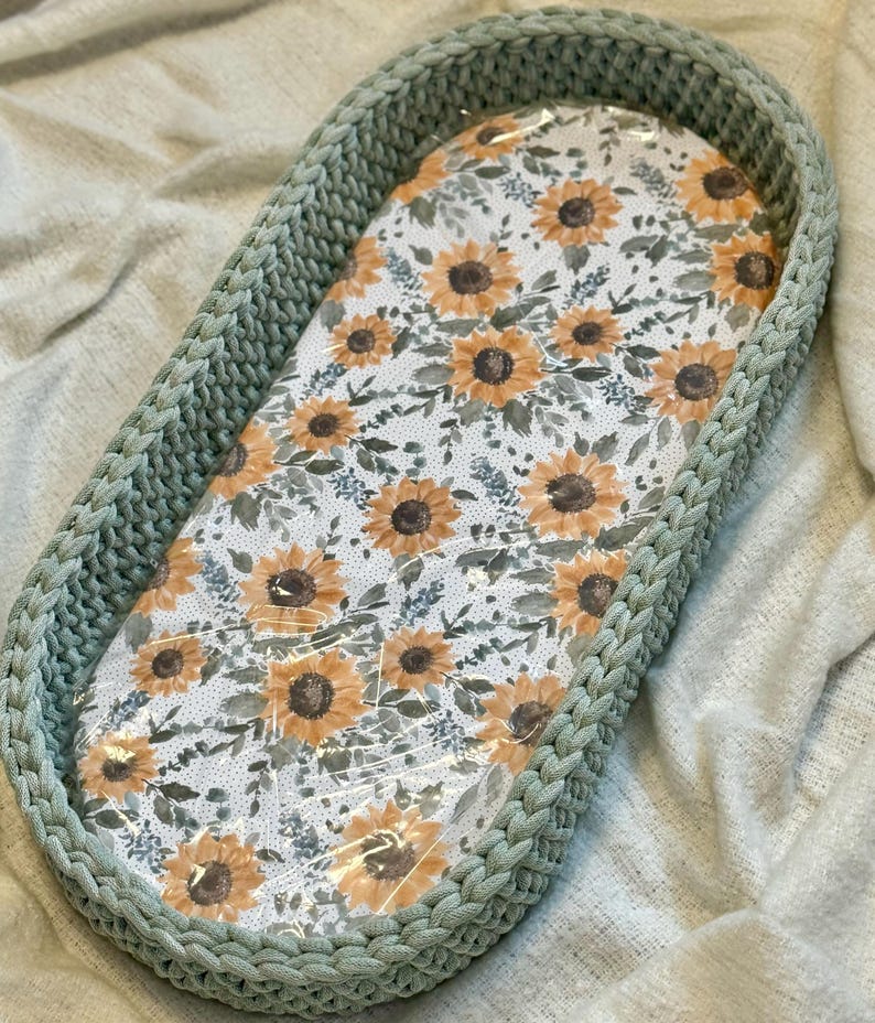 May include: An oval-shaped basket with thick, sage green crocheted walls and a flat base featuring a white fabric liner printed with a repeating pattern of yellow sunflowers, brown centers, and green foliage, covered by a clear plastic film.