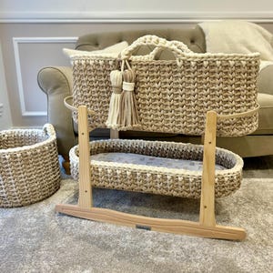May include: A woven bassinet with a natural beige and white color scheme. The bassinet is on a wooden rocking stand. There is a smaller woven basket in the background.