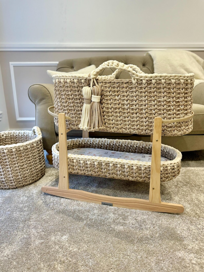 May include: A woven bassinet with a natural beige and white color scheme. The bassinet is on a wooden rocking stand. There is a smaller woven basket in the background.