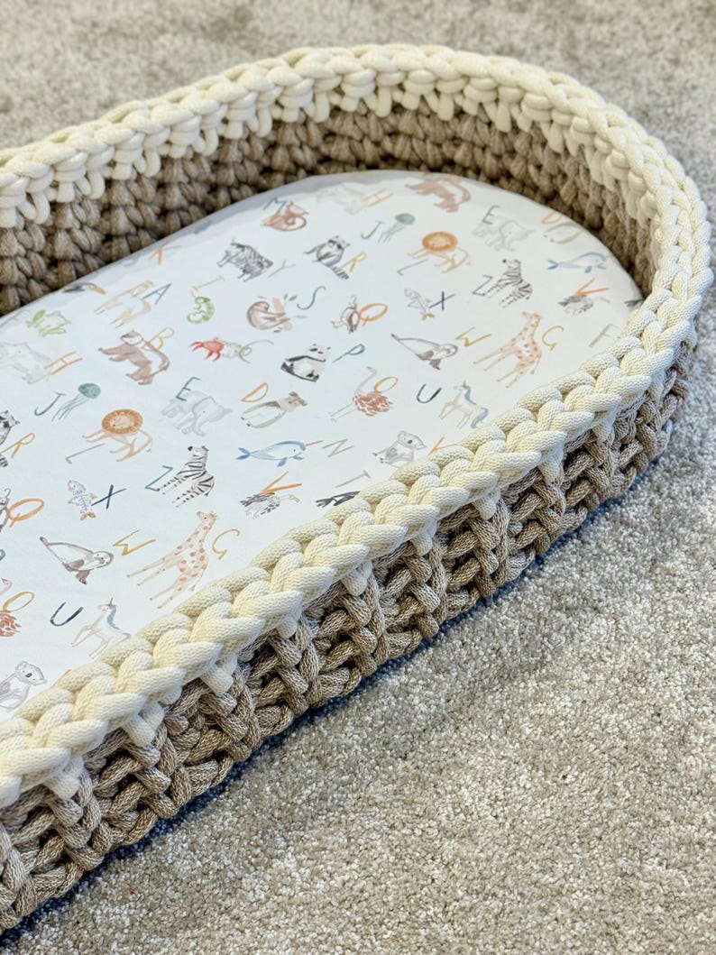 May include: A beige and white crocheted oval-shaped basket with a white fabric liner featuring a pattern of animals and letters.