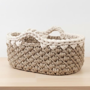 May include: A hand-crocheted basket in a neutral color palette. The basket features a light beige trim and handle, contrasting with the darker beige body. The oval-shaped basket is made of thick yarn, showcasing a textured, woven pattern.