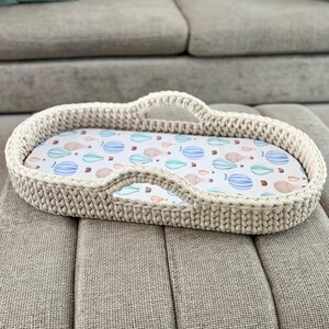 May include: An oval-shaped, cream-colored crocheted basket with two integrated handles. The interior features a white fabric liner decorated with a repeating pattern of small, colorful hot air balloons in shades of blue, green, and tan.