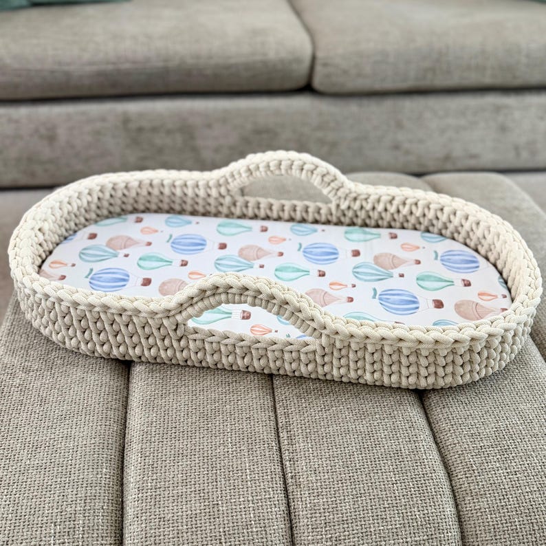 May include: An oval-shaped, cream-colored crocheted basket with two integrated handles. The interior features a white fabric liner decorated with a repeating pattern of small, colorful hot air balloons in shades of blue, green, and tan.