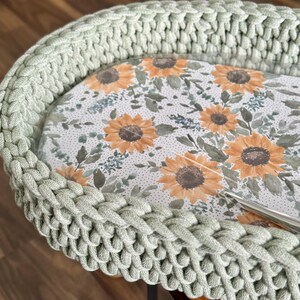 May include: A sage green crocheted basket with a thick, braided rim. The interior base features a printed pattern of yellow sunflowers, green leaves, and small dark dots on a white background. A silver metal crochet hook rests on the surface.