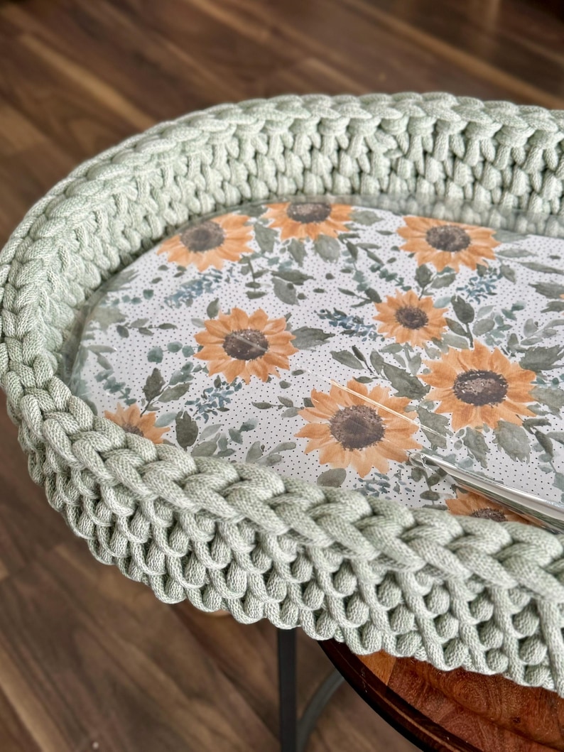 May include: A sage green crocheted basket with a thick, braided rim. The interior base features a printed pattern of yellow sunflowers, green leaves, and small dark dots on a white background. A silver metal crochet hook rests on the surface.