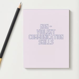 Op de afbeelding: Een lichtpaars notitieblok met de tekst "NON - VIOLENT COMMUNICATION SKILLS" in blauw. Een zwarte potlood met een gouden punt ligt diagonaal over de linkerbovenhoek van het notitieblok.