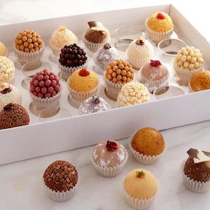May include: A white box filled with an assortment of colorful, bite-sized treats. The treats are round and covered in various toppings, including sprinkles, coconut flakes, and chocolate. Some are topped with a cherry or a piece of chocolate. The treats are in individual paper cups.