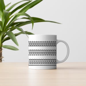 May include: White ceramic mug featuring a series of black geometric patterns. The designs include lines, zigzags, and wave-like shapes. The mug is set on a light-colored wooden surface.