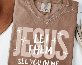 Op de afbeelding: Een bruin Comfort Colors T-shirt met de woorden "JESUS LET THEM SEE YOU IN ME" in witte en beige letters. Een zilveren ketting met een kleine hanger ligt op het shirt. Een bruine hoed is gedeeltelijk zichtbaar.