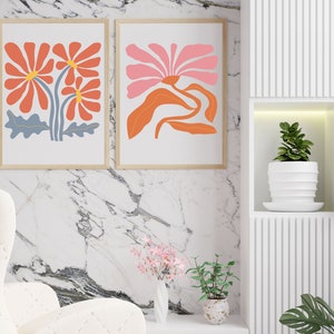 May include: Two framed prints of abstract floral designs. The print on the left features three orange flowers with blue stems and leaves. The print on the right features a single pink flower with orange leaves and stems.