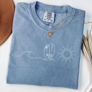 Puede incluir: Una camiseta azul Comfort Colors con un gráfico blanco de una ola, dos tablas de surf y un sol. Un collar de oro con un colgante está drapeado sobre la camisa. La camisa es talla S-P-CH.