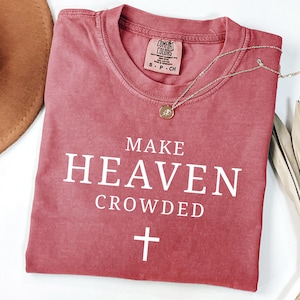 May include: A dusty rose Comfort Colors t-shirt with the phrase "MAKE HEAVEN CROWDED" and a cross graphic in white. A silver necklace with a pendant rests on the shirt. The shirt has a classic crew neck.
