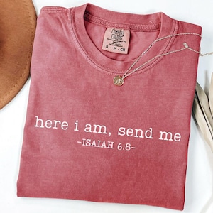 Op de afbeelding: Een stoffig roze Comfort Colors T-shirt met de witte tekst "here i am, send me - ISAIAH 6:8-". Een zilveren ketting met een kleine hanger ligt op het shirt. Het shirt is opgevouwen en tentoongesteld op een wit oppervlak.