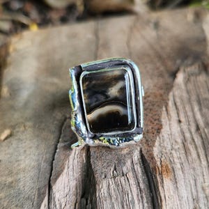 Pode incluir: Anel de prata artesanal com uma pedra retangular preta e branca. A pedra está engastada numa moldura de prata texturizada e detalhada. O anel é exibido numa superfície de madeira envelhecida.