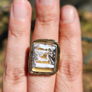 May include: A rectangular ring featuring a polished stone inlay with horizontal bands of white, translucent tan, and dark brown dendritic patterns. The stone is set within a thick, textured gold-toned metal bezel that frames the rectangular shape.