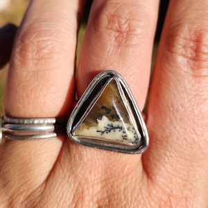 May include: A triangular ring featuring a polished dendritic agate stone with black fern-like inclusions set in a silver bezel. The stone displays a cream and tan background with dark branching patterns. A plain silver band is visible on the finger.