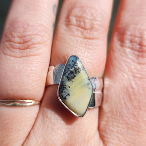 May include: A silver ring featuring a polished, kite-shaped dendritic opal gemstone. The stone displays a creamy yellow base color with dark, fern-like black inclusions branching across the surface. The metal band has a smooth, reflective finish.
