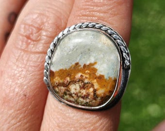Op de afbeelding: Een zilveren ring met een grote, onregelmatig gevormde steen. De steen heeft een mix van kleuren, waaronder lichtgroen, bruin en wit. De ring heeft een gevlochten zilveren band rond de steen.